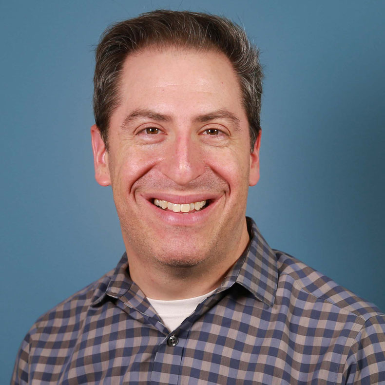 Evan J. Waldheter Portrait photo of man smiling, with short light brown hair wearing a checked shirt