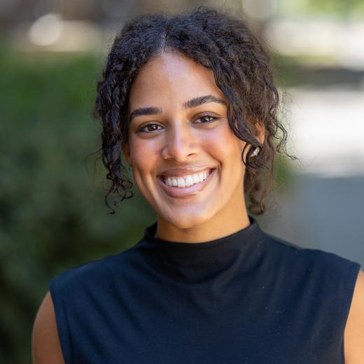 Gabriela Gonzalez, PsyD Portrait photo of female smiling with curly brown hair, dark blue sleeveless top, with blurred background