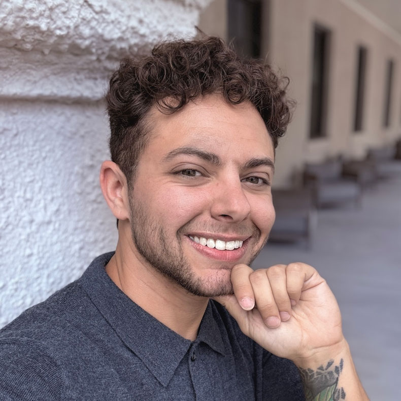 Parker Zyndorf, PsyD Portrait photo of smiling male, with hand to chin, curly brown hair, gray shirt, with a concrete column behind him and a blurred background