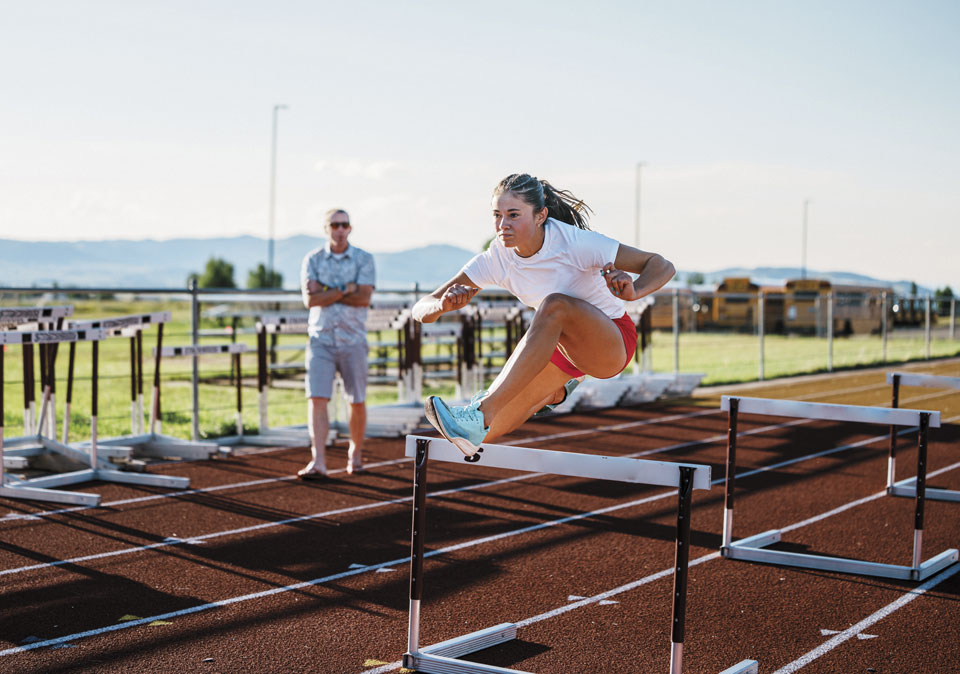 female athlete jumping hurdles with field and mountains in background and coach watching with arms crossed