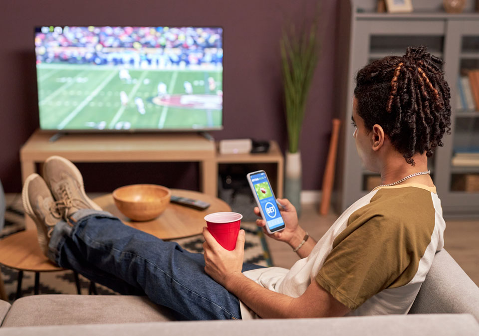 young man sitting on couch watching football game holding smart phone with a betting app showing 