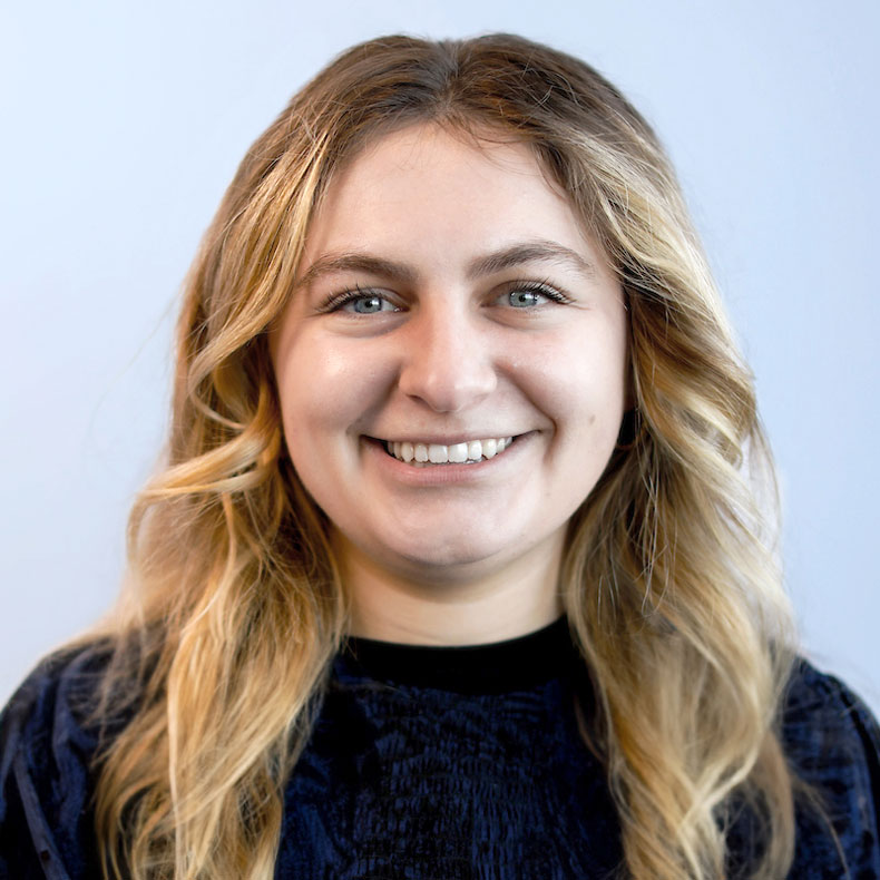 Headshot of woman smiling with blond hair and dark shirt on a light blue background