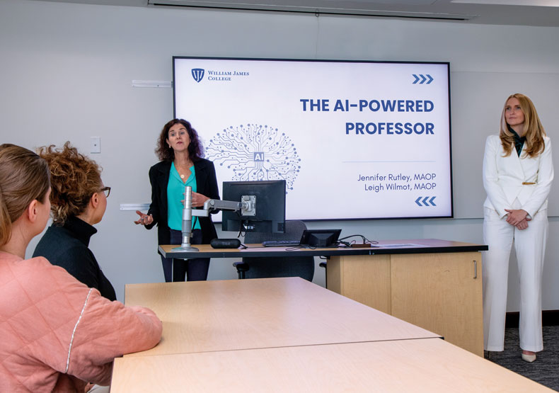 Two women standing in front of large display screen talking to others sitting at a table