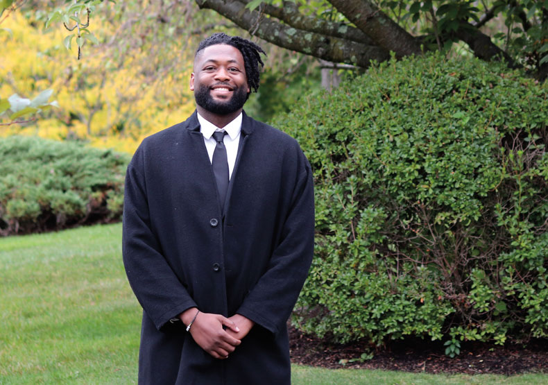 Black man in coat and tie smiling outside in front of large bush and tree