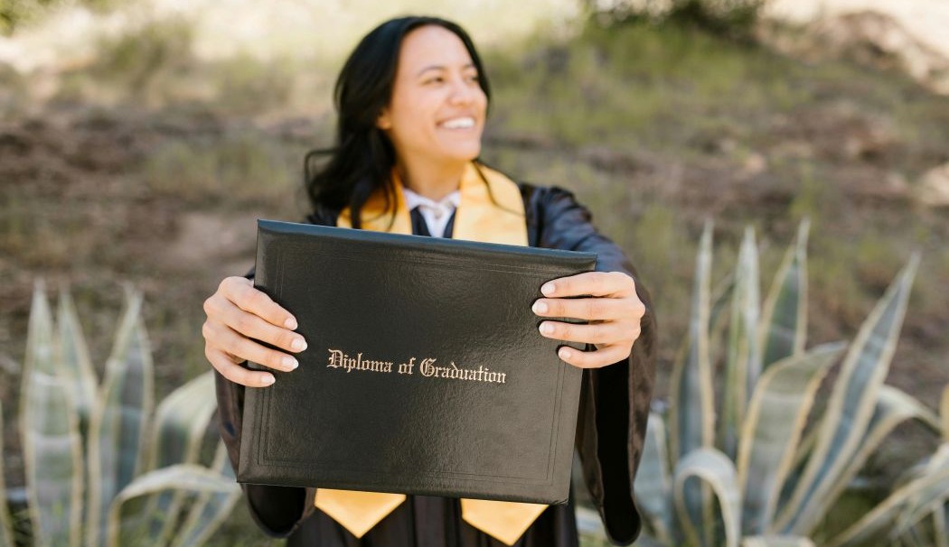 Photo of woman in academic regalia holding out a diploma in the foreground.