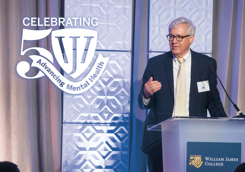 man in suit speaks from podium with light purple background and white logo in top left corner