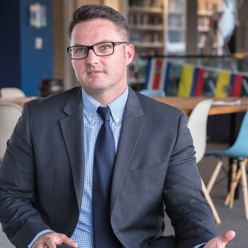 man in suit and glasses talking with bookshelves in the background