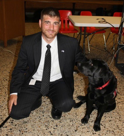 Kevin Lambert kneeling on the ground in a suit next to Ronnie, a black lab.