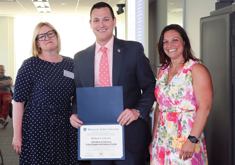 Man holding diploma, with two woman on either side smiling in classroom setting