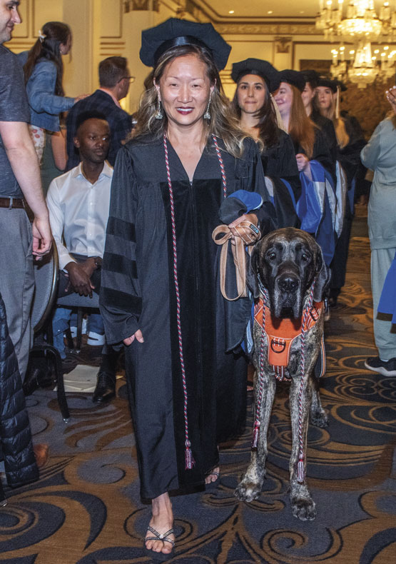 woman walking down aisle in commencement regalia, with service dog beside her