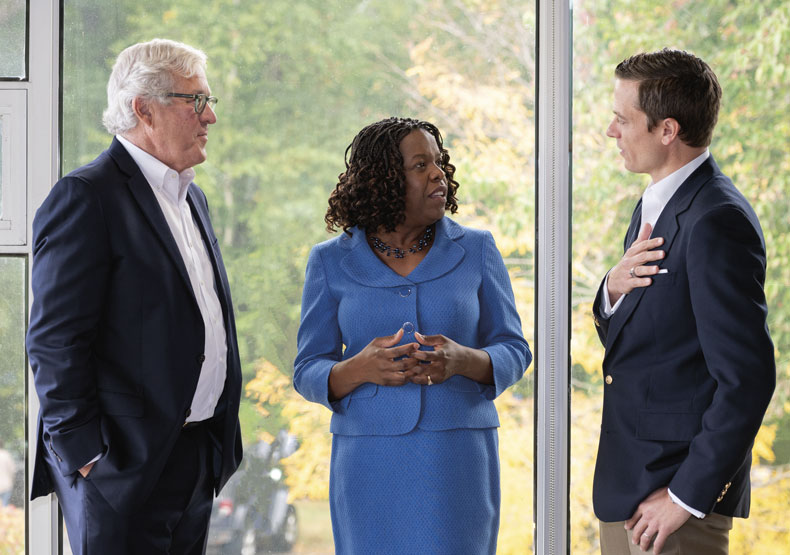 three people talking in front of large windows with trees in background