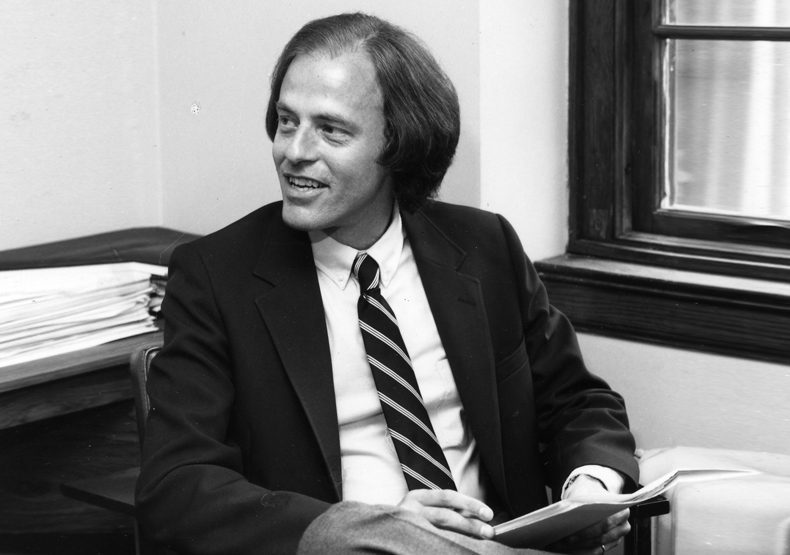 Black and white photo of man in suit and tie, sitting with papers in hands