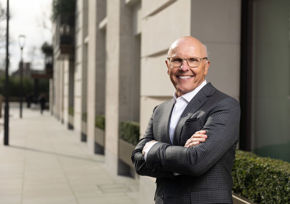 Man outside city building, smiling, wearing glasses, and in a dark suit with arms crossed