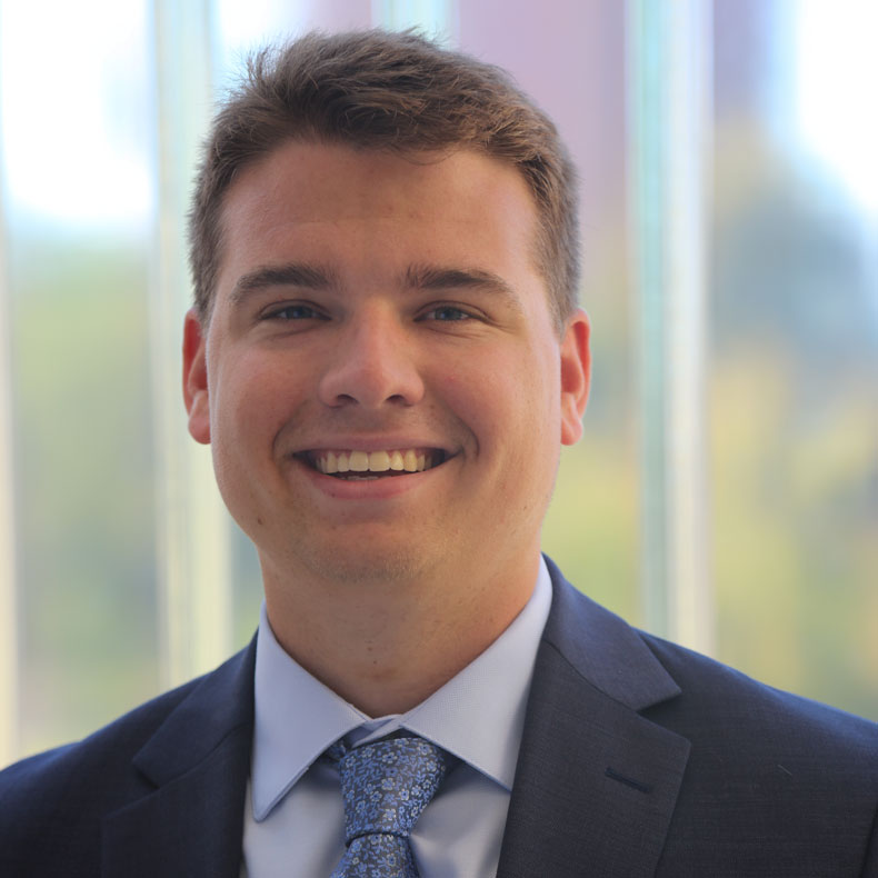 Headshot of smiling white male, with short light brown hair, in blue suit and tie with short brown hair with large window to outside behind him