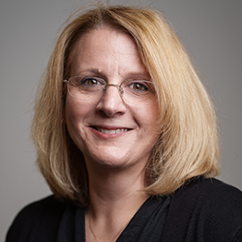 Portrait photo of woman smiling, with shoulder-length strawberry blonde hair, wearing glasses, in a black top and a neutral background